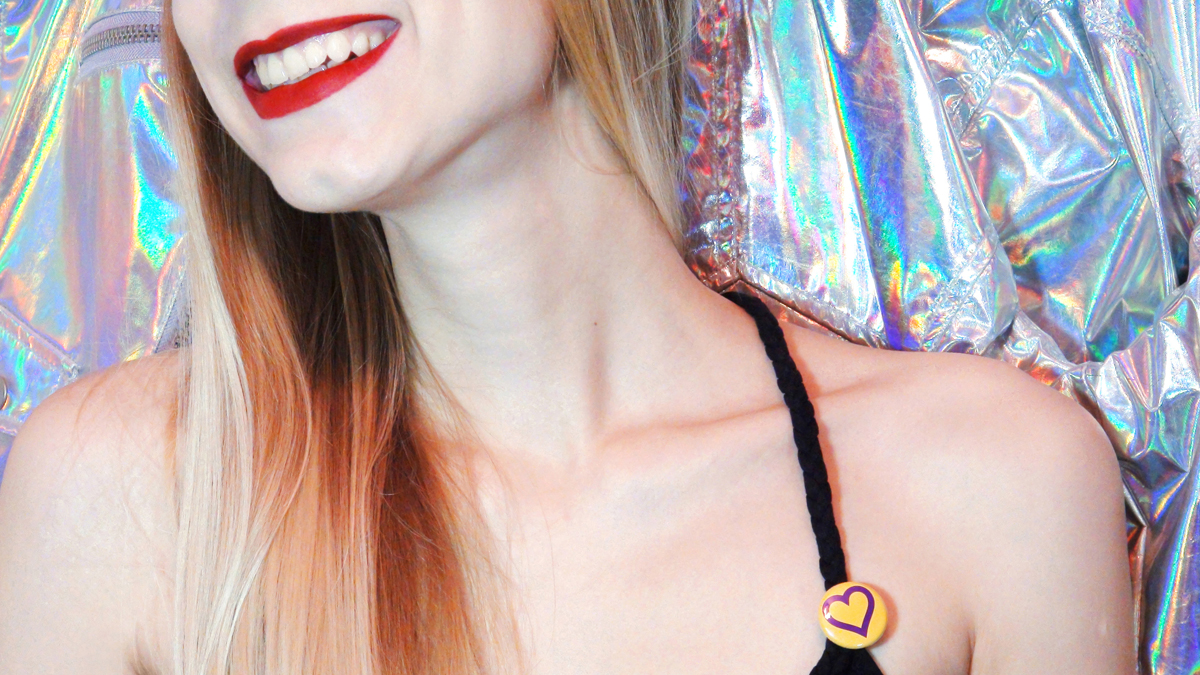 Image of Irene, who discovered she was intersex without knowing it. The shot shows only her mouth, smiling in red lipstick. She wears a small intersex pin on her tank top strap, and stands in front of a colorful cloth background.