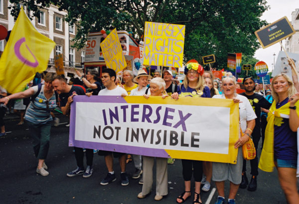 A crowd of intersex people holding signs at a LGBTQIA Pride parade, five in front row hold a large banner with text 'Intersex, Not Invisible'