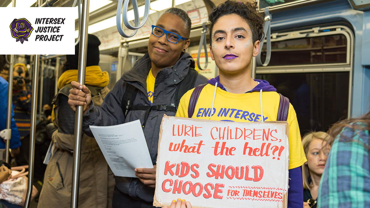 Intersex Justice Project co-founders Saifa and Pidgeon smile while holding protest signs in a Chicago train, on their way to lead a protest to make Lurie Children's Hospital end intersex surgery.