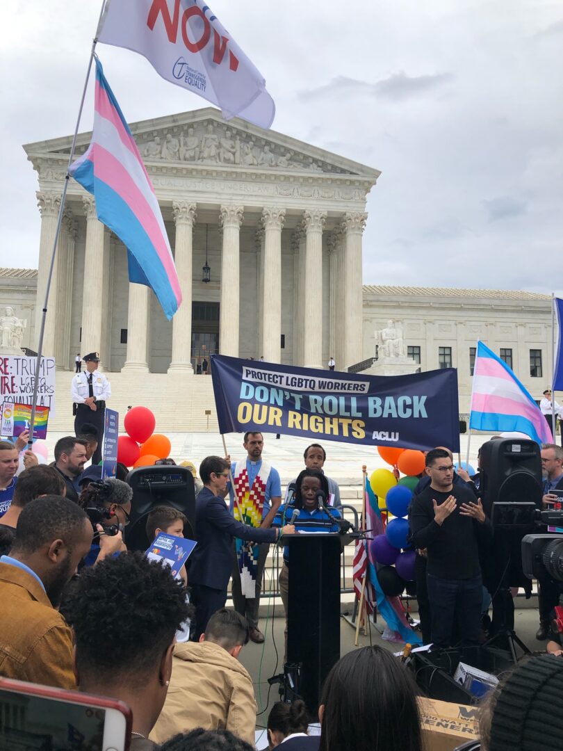 Bria, a Black masculine-presenting intersex person, stands at a small podium on the steps of the Supreme Court, speaking, surrounded by crowds of reporters and waving transgender flags.