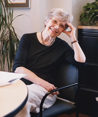 Lois, an older white woman wearing a black sweater and glasses with short white hair, smiles while sitting at her desk, hand holding her face.