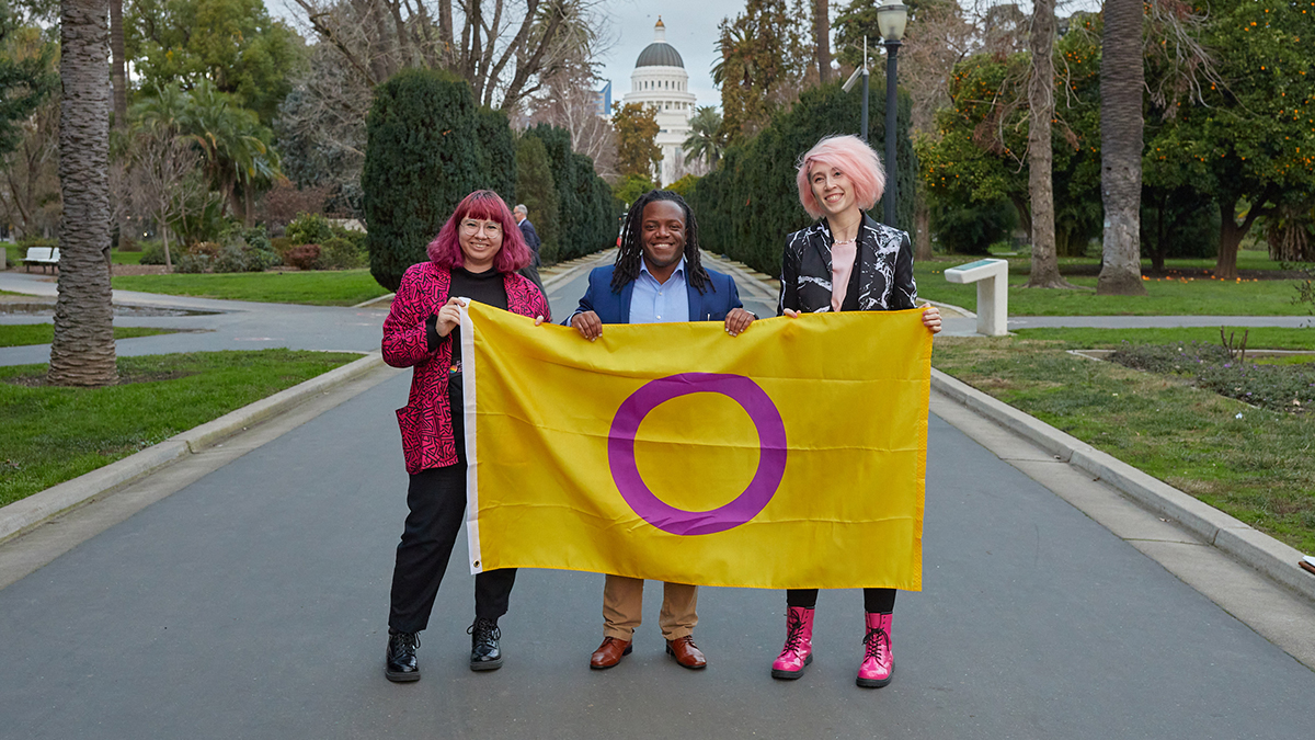 Three young intersex people stand smiling holding an intersex flag in support of SB 225. The Sacramento capitol dome is seen in the background behind them.