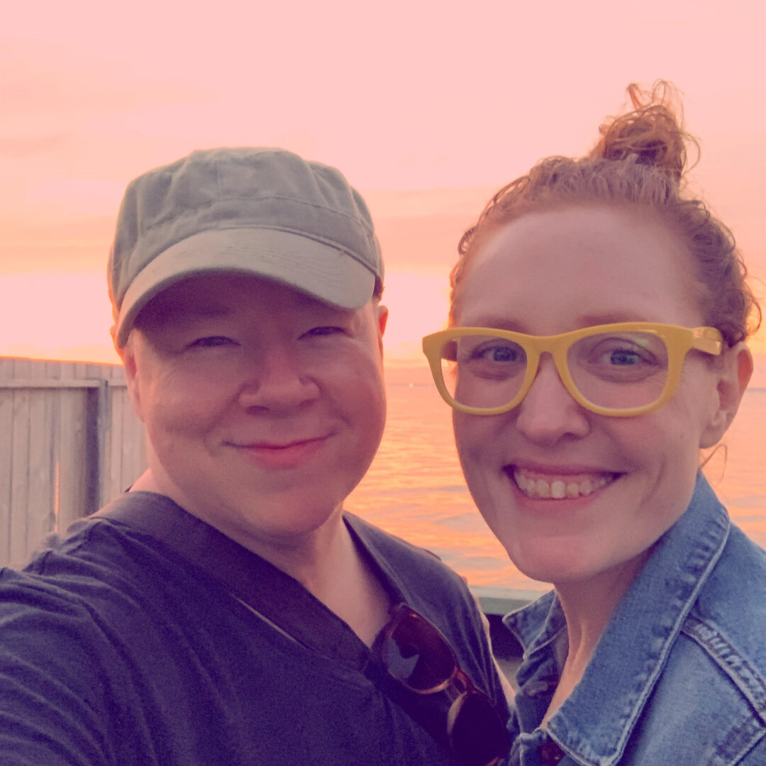 interACT's Executive Director Erika and Katie pose on Fire Island. Erika is a white intersex person wearing a hat and smiling widely at the camera, Katie is a white person with red hair in a bun and wears yellow-rimmed glasses, also smiling widely. The two are very close to each other with arms around each other.