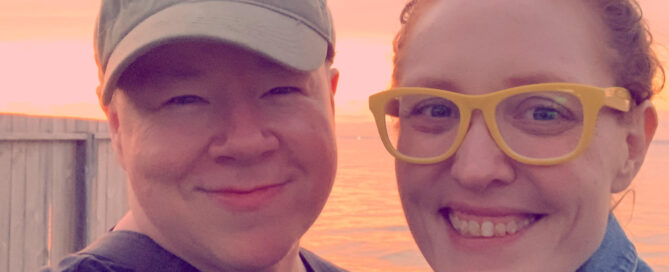 Erika and Katie pose on Fire Island. Erika is a white intersex person wearing a hat and smiling widely at the camera, Katie is a white person with red hair in a bun and wears yellow-rimmed glasses, also smiling widely. The two are very close to each other with arms around each other.