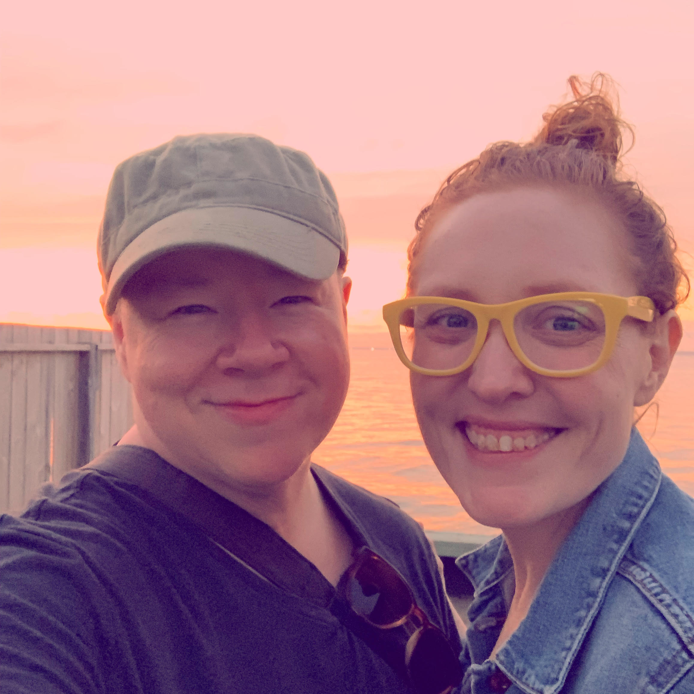 Erika and Katie pose on Fire Island. Erika is a white intersex person wearing a hat and smiling widely at the camera, Katie is a white person with red hair in a bun and wears yellow-rimmed glasses, also smiling widely. The two are very close to each other with arms around each other.