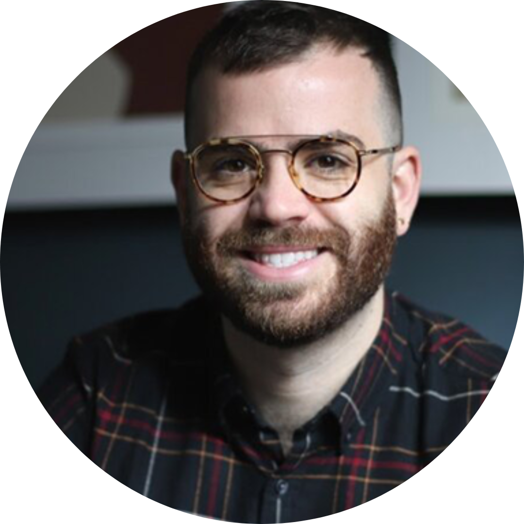 Aaron is a cisgender, white, queer man with short, dark hair and thinly rimmed glasses. He has a beard and wears a flannel shirt and smiles widely at the camera.