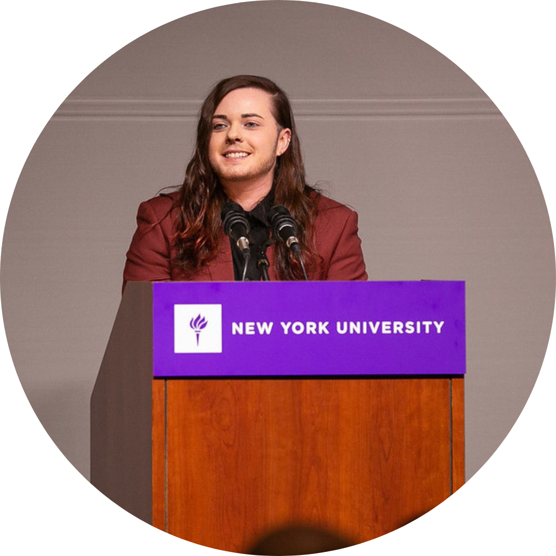 Axel is a light-skinned nonbinary person with brown, long hair and facial hair. They stand at an NYU podium smiling.