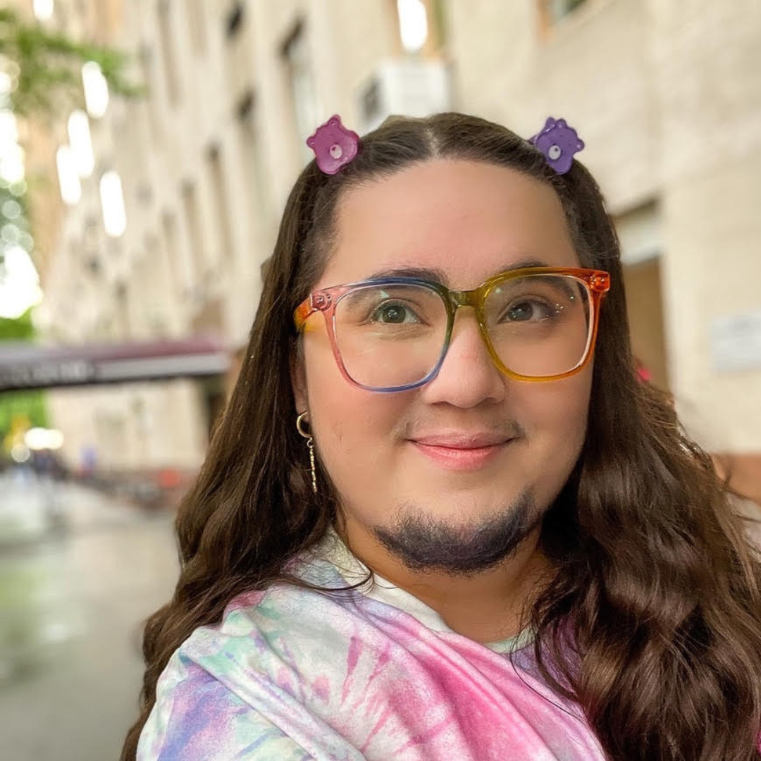 Liat, a person with long hair in butterfly clips, rainbow glasses who is smiling at the camera.