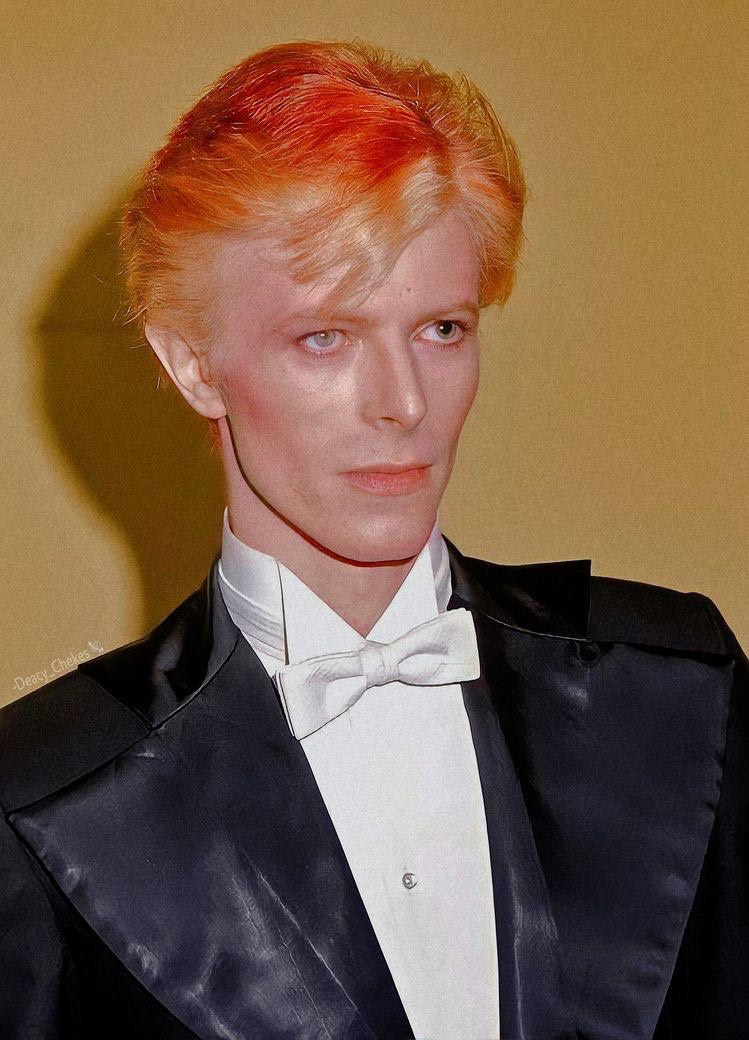 A photograph of David Bowie at the 1975 Grammys. He is looking to the right, and is dressed in a tuxedo blazer with large lapels. He has a white bowtie, and red-orange dyed hair with blonde roots.