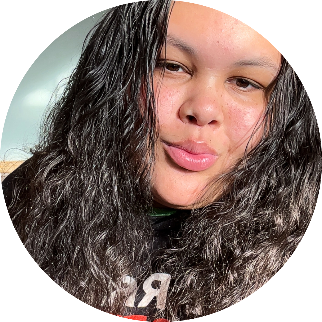 Justin is an Asian American intersex person. He has short, dark hair and wears a jean jacket over a white t-shirt. He smiles widely in front of a city background of large buildings.