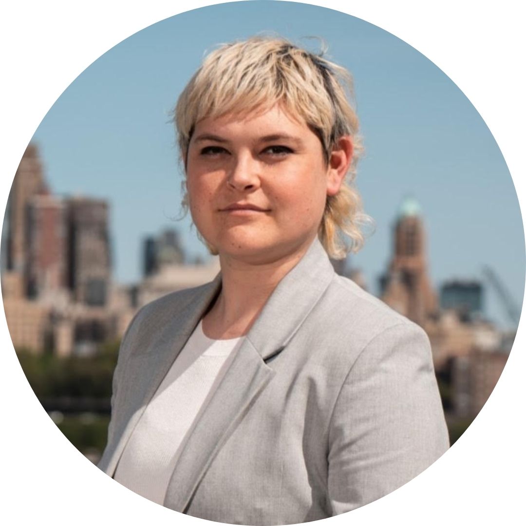 Shana is a white woman with short, bleach-blonde hair and dark eyes wearing a grey suit. She stands against a city background.